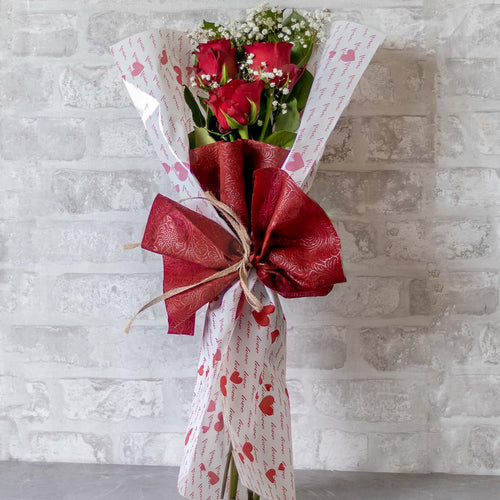 Bouquet of red roses wrapped in white paper with red floral patterns against a light gray brick wall.