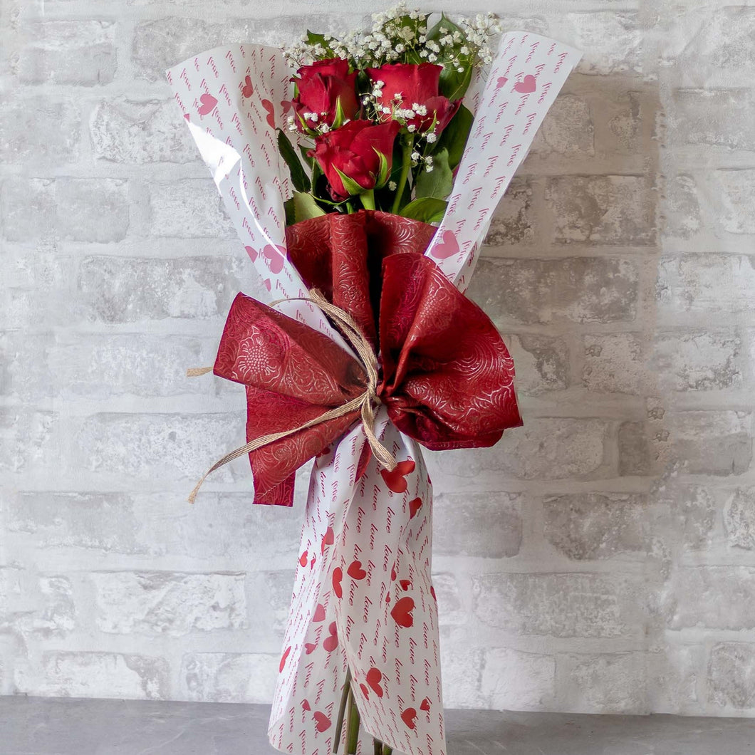 Bouquet of red roses wrapped in white paper with red floral patterns against a light gray brick wall.