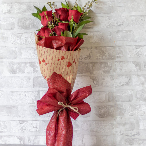 Bouquet of red roses wrapped in red paper with a bow against a light gray brick wall.