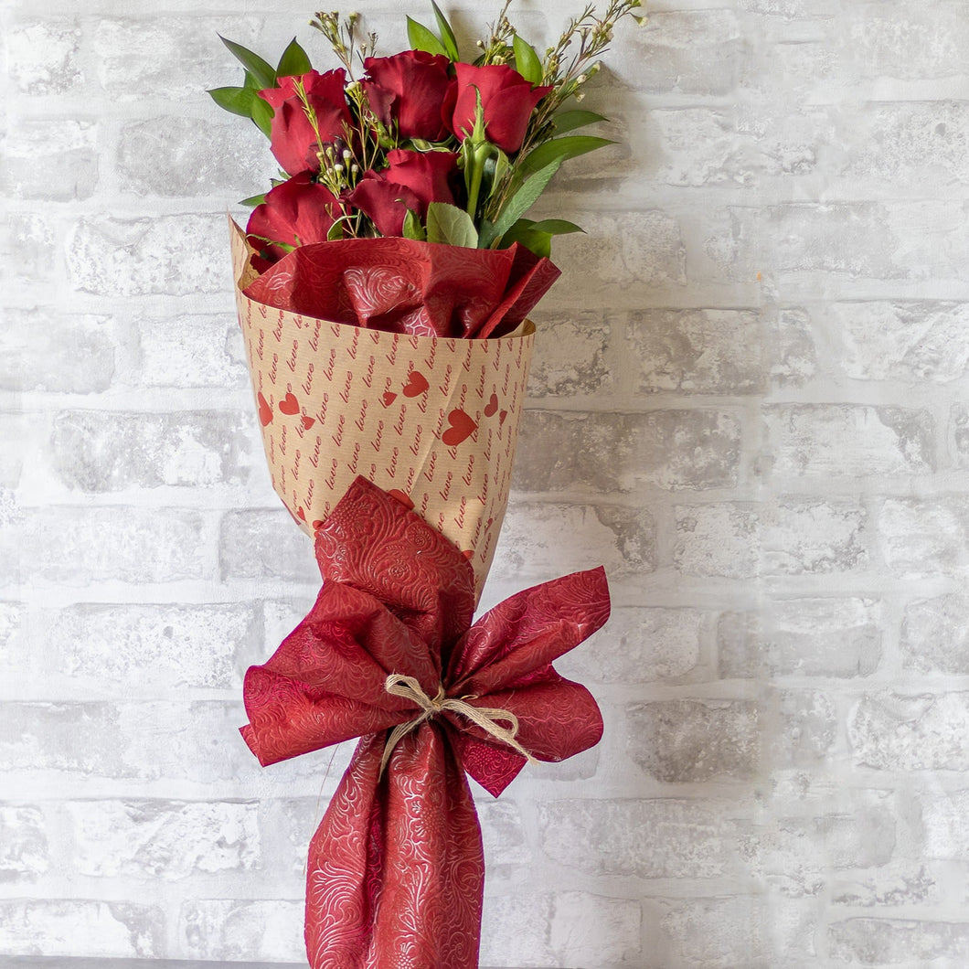 Bouquet of red roses wrapped in red paper with a bow against a light gray brick wall.