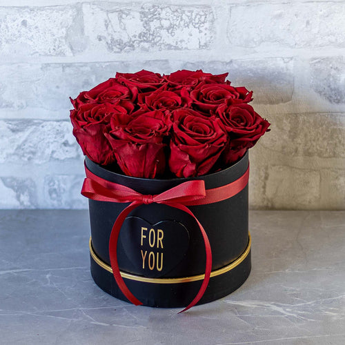 Black box with red roses and 'FOR YOU' ribbon against a light gray brick wall.