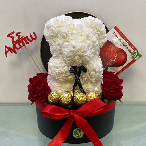 White teddy bear made of flowers in a black box with red ribbon, surrounded by red roses and a small black dog figurine on a light gray surface.