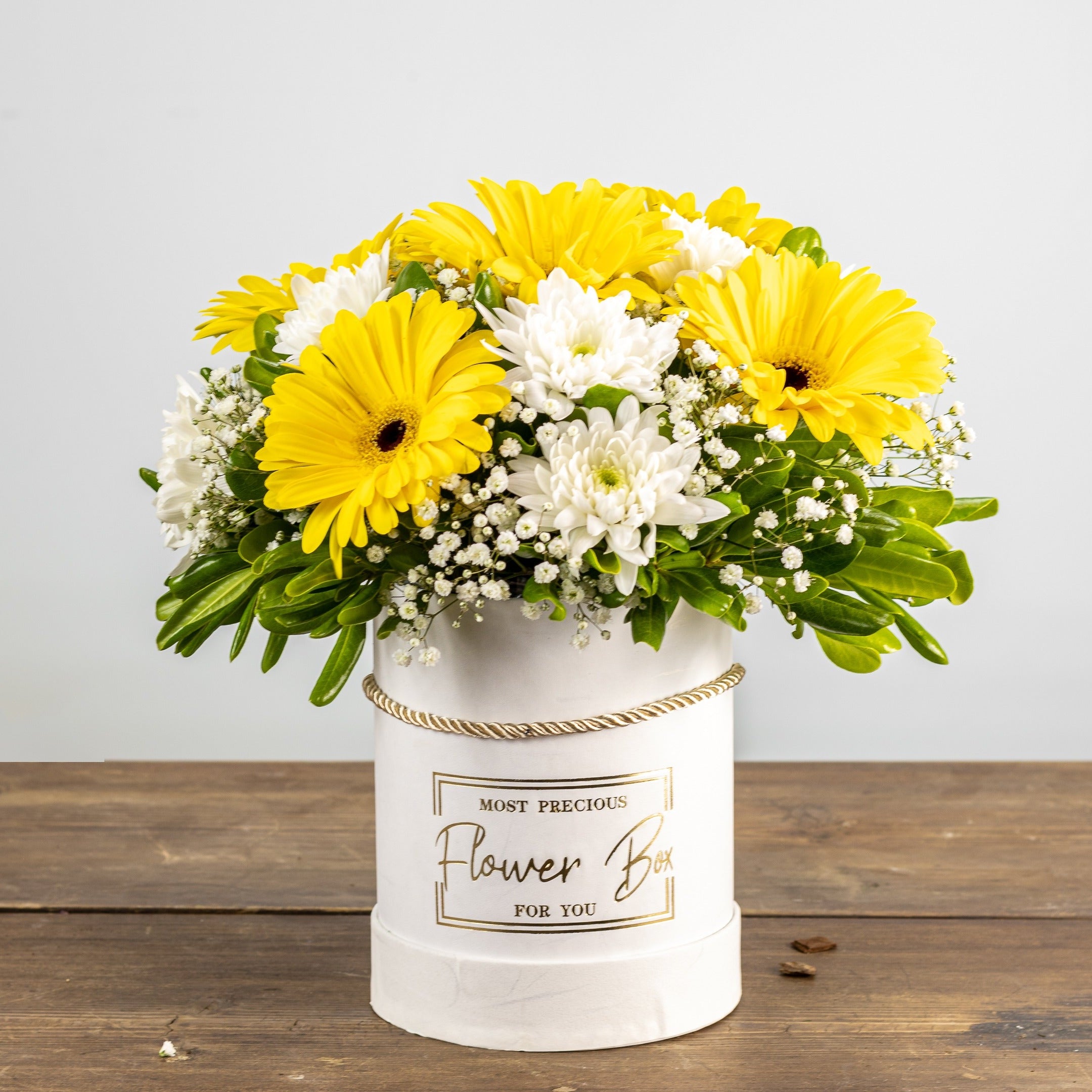 Bouquet of yellow and white flowers in a white container on a wooden surface with a light gray background.