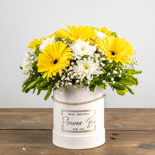 Bouquet of yellow and white flowers in a white container on a wooden surface with a light gray background.