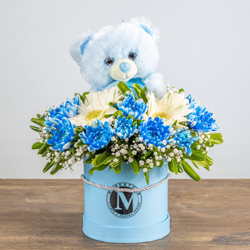 Floral arrangement with blue flowers and a teddy bear in a light blue container on a wooden surface.
