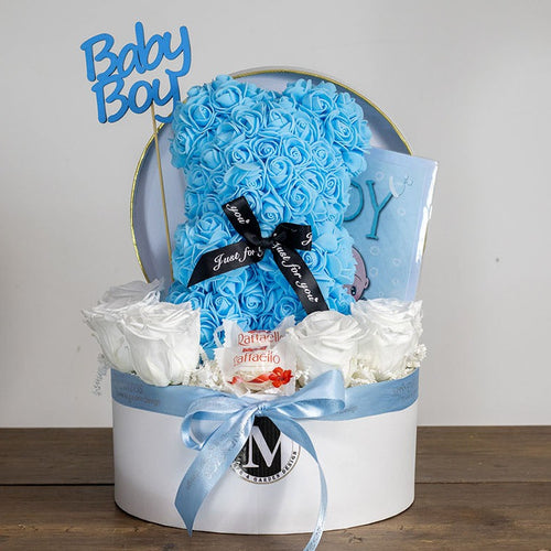 Gift basket with blue teddy bear, white flowers, and 'Baby Boy' sign on a wooden surface.