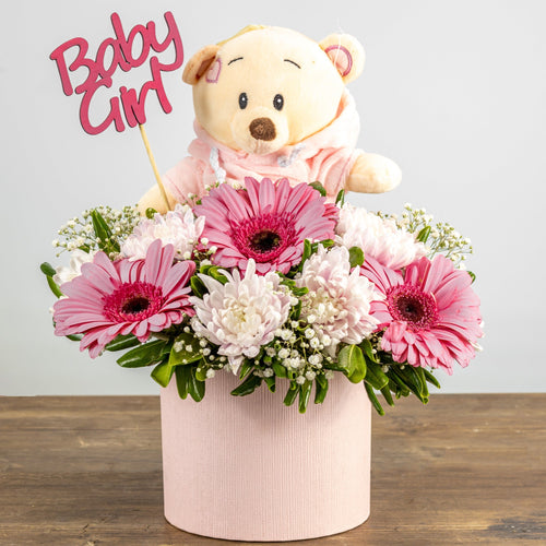 Floral arrangement with pink flowers and a teddy bear wearing a 'Baby Girl' outfit on a wooden surface.