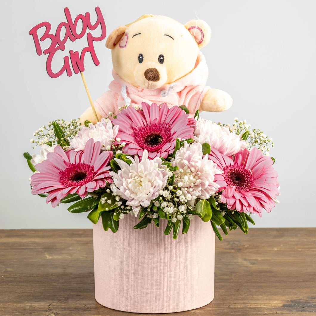 Floral arrangement with pink flowers and a teddy bear wearing a 'Baby Girl' outfit on a wooden surface.