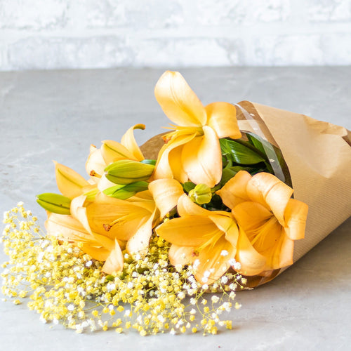 Bouquet of yellow flowers wrapped in brown paper on a light gray surface with a white brick wall background.