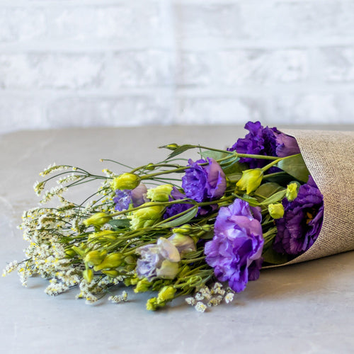 Bouquet of purple and yellow flowers wrapped in burlap on a light surface with a white brick wall background.