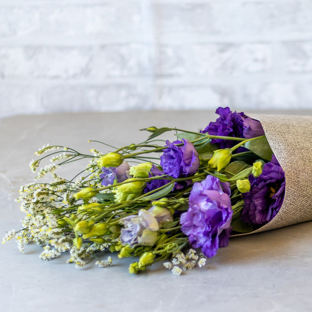 Bouquet of purple and yellow flowers wrapped in burlap on a light surface with a white brick wall background.