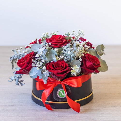 Floral arrangement with red roses and greenery in a black box with a red ribbon on a light background