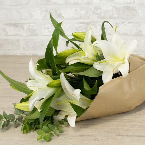Bouquet of white lilies wrapped in brown paper on a wooden surface with a light gray tiled wall background.