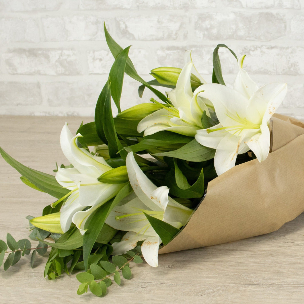 Bouquet of white lilies wrapped in brown paper on a wooden surface with a light gray tiled wall background.